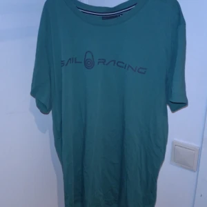 Grön t-shirt från Sail Racing - Säljer en grön t-shirt från Sail Racing i bra skick. Den har en normal passform och är kortärmad, perfekt för vardagsbruk eller träning. Loggan är tryckt på framsidan och baksidan. Skön och stilren design! Kan mötas Trelleborg eller Malmö 