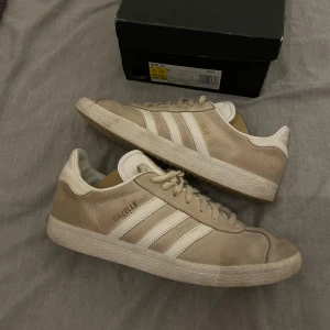 Beige/vita Adidas Gazelle Sneakers - Säljer ett par snygga beige/vita Adidas Gazelle sneakers i mocka i storlek 42 2/3. De har den klassiska tre-randiga designen och en bekväm sula. Perfekta för både vardag och fest! Skorna är i mycket bra skick och redo för nya äventyr.🥂