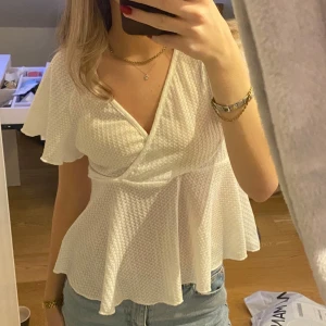 Vit omlottopp - Säljer en superfin vit omlottopp med volangdetaljer. Toppen har en v-ringad design och korta ärmar, perfekt för sommardagar. Materialet är lätt och luftigt, vilket gör den bekväm att bära. Passar perfekt till jeans eller kjol för en avslappnad men stilren look.