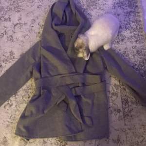 Grå omlottjacka med bälte - Säljer en superfin grå omlottjacka med bälte. Perfekt för höst och vår! Jackan har långa ärmar och en mysig huva. Den är i bra skick och passar perfekt för en casual look. Perfekt för att hålla sig varm och stilren samtidigt!