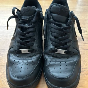 Svarta Nike Air Force sneakers - Säljer ett par svarta Nike Air Force sneakers i bra skick. De är tillverkade i skinn och har klassisk snörning. Perfekta för både vardag och fest. Storlek är inte angiven, men de ser ut att passa en normal passform. Skorna har en stilren design med perforerade detaljer på ovansidan.