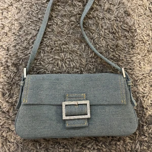 Jeansväska med glittrigt spänne - Säljer en snygg axelväska i jeansmaterial med ett glittrigt spänne framtill. Väskan har en praktisk magnetknäppning och en justerbar axelrem. 🩵🩵