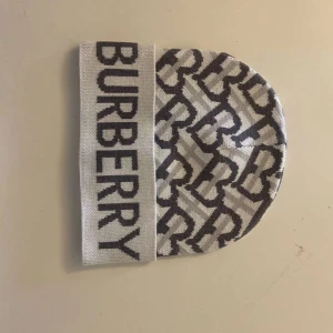 Vit och grå mössa från Burberry - Snygg och stilren mössa från Burberry i vitt och brunt. Den har ett coolt mönster med Burberry-loggan och är perfekt för att hålla dig varm med stil. Passar perfekt till både vardag och lite finare tillfällen. Ett måste för modeälskaren!