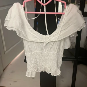 Vit offshoulder topp - Säljer en söt vit offshoulder topp med volangdetaljer. Toppen är croppad och har en smockad midja som ger en fin passform. Perfekt för sommarens fester eller en dag på stan. Den är i mycket bra skick och redo för en ny ägare!
