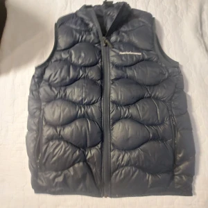 Blå dunväst från Peak Performance - Säljer en snygg blå dunväst från Peak Performance i storlek 160. Västen har en dragkedja framtill och är perfekt för kyliga dagar. Den är quiltad och ger ett stilrent intryck. Passar både för höst och vår. Perfekt för dig som vill hålla värmen med stil!