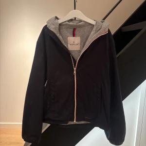 Marinblå  jacka med huva från Moncler - Säljer en snygg marinblå jacka från Moncler i väldig bra skick. Den har en grå huva och dragkedja framtill. Perfekt för höst och vår! Jackan har långa ärmar och en stilren design med Moncler-logga på ärmen.