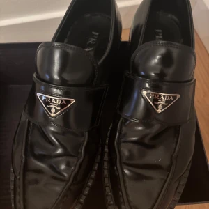 Prada Loafers - Säljer ett par eleganta svarta loafers från Prada i skinn. De har en stilren design med Pradas logotyp på ovansidan. Perfekta för både formella och avslappnade tillfällen. Skorna är i mycket bra skick och kommer i originalförpackning samt kvitto. Nypris ca 10000 kr