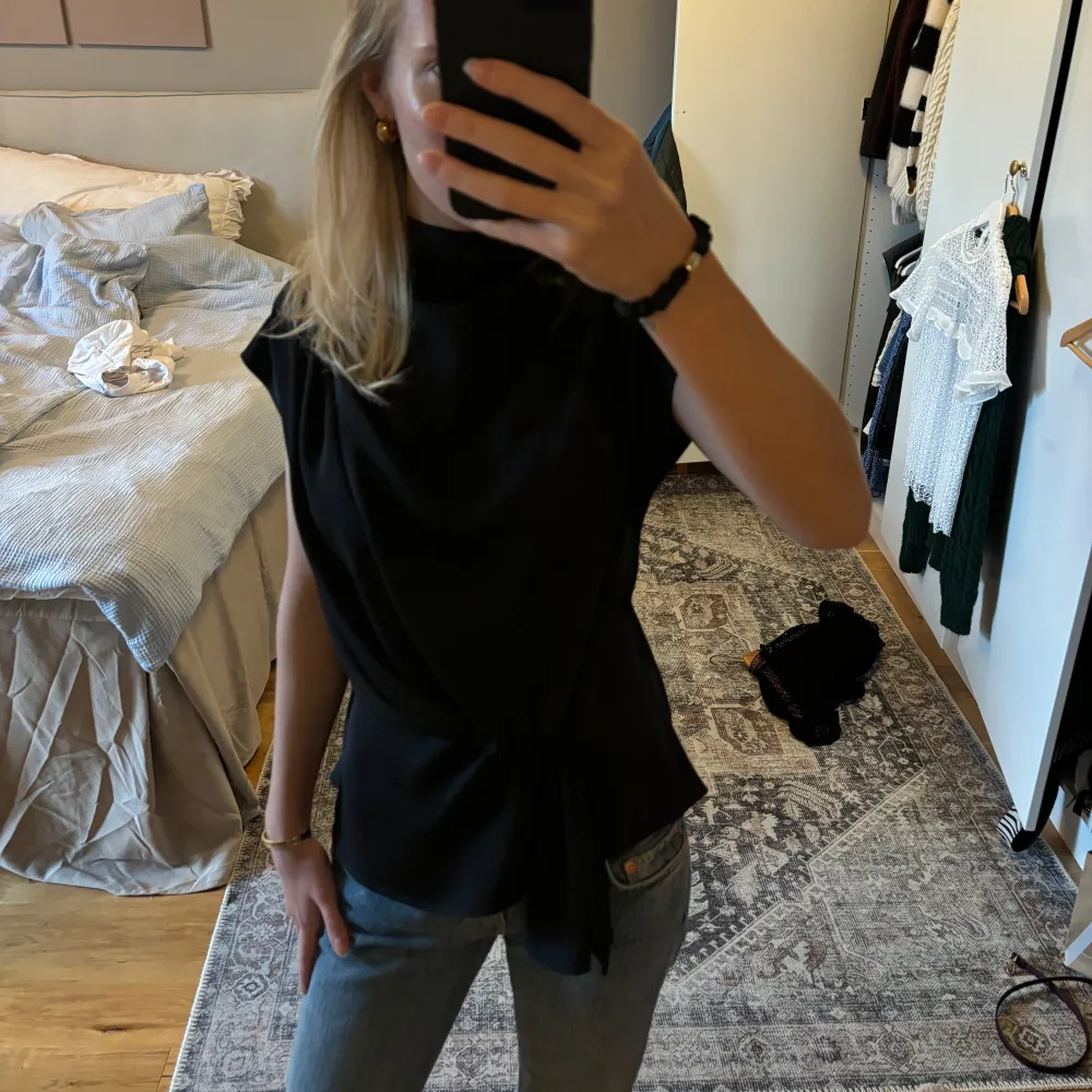 Säljer en stilren svart topp med knytning i midjan. Den är ärmlös och har en elegant, draperad design som passar perfekt för både vardag och fest. Materialet är satin och känns mjukt och bekvämt mot huden. Perfekt för att styla med jeans eller kjol för en chic look!. Toppar.