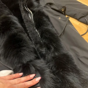 Svart parkajacka med pälsdetaljer - Säljer en snygg svart parkajacka S. Jackan har lyxiga pälsdetaljer längs kragen och ärmarna, perfekt för kalla vinterdagar. Den är i mycket bra skick och har en stilren design med knappar och dragkedja. Passar perfekt för både vardag och festliga tillfällen. Passar till både XS/S