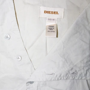 Vit/ ljusgrå skjorta från Diesel - Säljer en stilren vit/ ljusgrå skjorta från Diesel i storlek XS. Skjortan är tillverkad i 100% bomull och har en unik asymmetrisk knäppning framtill. Den har långa ärmar och en hög krage, perfekt för både vardag och fest. Skjortan är i mycket bra skick och redo för nya äventyr!