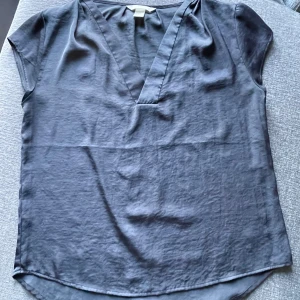Blå blus från H&M - Säljer en stilren grå blus från H&M  i storlek M. Blusen har en v-ringad design och korta ärmar, perfekt för både vardag och fest. Materialet är silkigt, mjukt och bekvämt mot huden. 