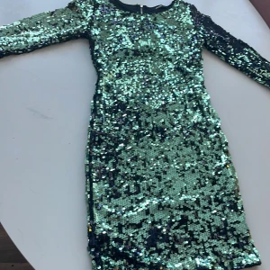 Grön paljettklänning med öppen rygg - Snygg grön paljettklänning med långärmad design och en cool öppen rygg. Perfekt för festliga tillfällen när du vill glittra och sticka ut. Klänningen har en dragkedja bak för enkel påklädning.
