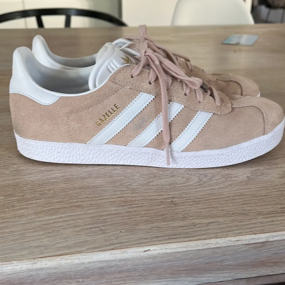 Säljer ett par snygga beige Adidas Gazelle sneakers i mocka. De har de klassiska vita ränderna på sidan och 'GAZELLE' i guldtext. Skorna är i bra skick och perfekta för både vardag och fest. De har en vit sula och snörning framtill.. Kengät.