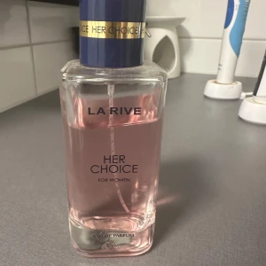 La Rive Her Choice Eau de Parfum - Säljer en elegant och feminin parfym från La Rive, 'Her Choice'. Flaskan är stilren och rektangulär med en ljusrosa vätska inuti. Korken är mörkblå med en guldfärgad detalj. Perfekt för dagligt bruk eller speciella tillfällen.