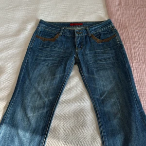 Blå jeans med broderade detaljer - Säljer ett par snygga blå jeans från Bonny Star. De har coola broderade detaljer vid fickorna och en unik patch på baksidan med texten 'Pick your poison'. Jeansen har en klassisk femficksdesign och en bekväm passform. De är low waist och bootcut. Perfekta för en avslappnad stil eller festlig dag! Inga defekter och knappt använda. Säljer pga att de inte passar mig.