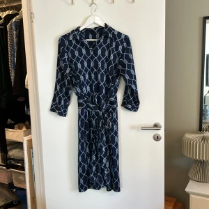 Mönstrad klänning från Gina Tricot - Säljer en snygg mörkblå klänning från Gina Tricot med ett unikt mönster av ljusblå rep. Klänningen har trekvartsärmar och ett knytband i midjan för en fin passform. Perfekt för både vardag och fest!