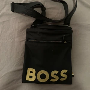 Boss Axelremsväska  - Stilig axelrems väska, justerbar axelremsväska så passar alla storlekar. Lite defekter som man Kan se på bilden på själva boss texten annars är väskan i super bra skick. Bara att höra av säg vid fler bilder/frågor!