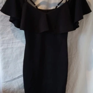 Svart klänning med volangdetaljer - Säljer en elegant svart klänning med volangdetaljer vid axlarna. Perfekt för festliga tillfällen! Klänningen har en tight passform och är axelbandslös med smala band för extra stöd.
