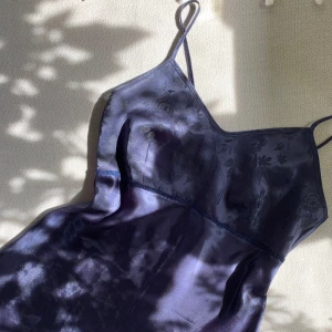 Mörkblå silkig klänning - Ett vackert nattlinne i mörkblått i silkigt material med ett blommigt mönster. Klänningen har tunna band med justerbar längd då de knyts baktill. Knappt använd men tyget har fått några små (knappt märkbara) repor framtill - se sista bilden.