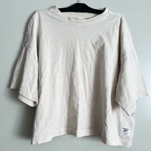 Beige t-shirt från Reebok - Säljer en snygg beige t-shirt från Reebok. Den är gjord av mjukt bomullsmaterial och har en avslappnad passform. Perfekt för en casual look eller träning. T-shirten har korta ärmar och en liten Reebok-logga på framsidan. Den är i nyskick och redo för nya äventyr!