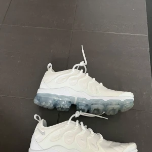 Vita Nike Air Vapormax - Säljer ett par vita Nike Air Vapormax med genomskinlig sula och snörning. Skorna har en modern design med synliga luftkuddar i sulan för extra komfort och stil.