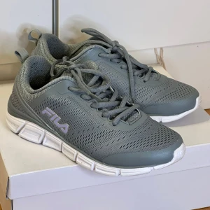Grå sneakers från Fila - Snygga grå sneakers från Fila med vit sula och snörning. Skorna har en sportig design med ventilerande meshmaterial och Fila-logga på sidan. Perfekta för en aktiv livsstil.