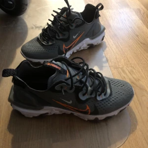 Nike sneakers (React Vision) storlek 41 - Snygga grå Nike sneakers med orange swoosh och svarta snören. Skorna har en modern design med vit sula och perforerat mönster för extra stil. Perfekta för en sportig look.  Storlek 41. Nypris: 1800. Bra skick