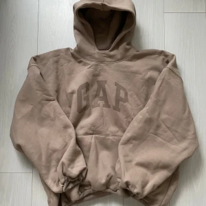 Beige hoodie från YZY x GAP - Säljer en beige hoodie från YZY x GAP med stor logga framtill och en vit fågeltryck på baksidan. Hoodien har en rymlig passform och en stor ficka framtill. Perfekt för en avslappnad stil.