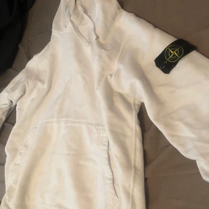 Vit hoodie från Stone Island - Säljer en stilren vit hoodie från Stone Island med deras ikoniska märke på ärmen. Tröjan har en klassisk design med huva och en stor ficka framtill. Perfekt för en avslappnad look. Pris kan diskuteras vid snabb affär