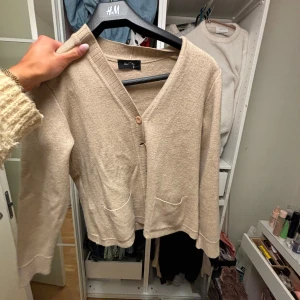 Beige kofta från H&M - Säljer en stilren beige kofta från H&M med v-ringning och knappar framtill. Koftan har långa ärmar och två små fickor fram. Perfekt för lager-på-lager under kyligare dagar.