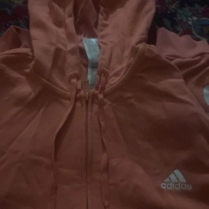 Pink hoodie från Adidas - Säljer en cool pink hoodie från Adidas med dragkedja och huva. Den har Adidas-loggan tryckt på bröstet och längs ena ärmen. Perfekt för en sportig look eller avslappnade dagar. 🧡