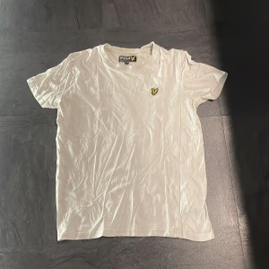 Vit t-shirt från Lyle & Scott - Säljer en klassisk vit t-shirt från Lyle & Scott med deras ikoniska gula logga broderad på bröstet. Perfekt för en stilren och enkel look. T-shirten är kortärmad och gjord i mjukt bomullsmaterial. Köpt på Lyle & Scott.