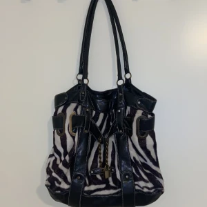 Zebramönstrad axelväska med kedjedetalj - Snygg axelväska med zebramönster och svarta skinnliknande detaljer. Väskan har en cool kedjedetalj framtill och metallspännen som ger en edgy look. 