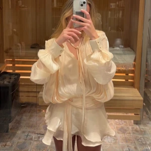 Vit klänning med volanger - Säljer en eleganta vit/champagnefärgad klänning med volanger och knytband i midjan. Aldrig använt nypris är 1800 kr.  Klänningen har långa ärmar med knappar vid ärmsluten och en v-ringad design. Den har shorts under i samma material som klänningen vilket är så skönt speciellt om man ska ha den till student eller liknande. 