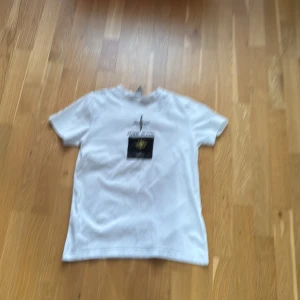 Vit t-shirt från Stone Island - Säljer en stilren vit t-shirt från Stone Island med ett broderat emblem på framsidan. T-shirten har korta ärmar och är tillverkad i mjukt bomullsmaterial. Perfekt för en avslappnad look.