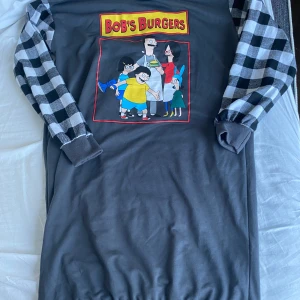 Grå Bob's Burgers tröja med rutiga ärmar - Säljer en cool grå tröja med Bob's Burgers-tryck på framsidan. Tröjan har svartvita rutiga ärmar och en avslappnad passform. Perfekt för fans av serien! 🖤