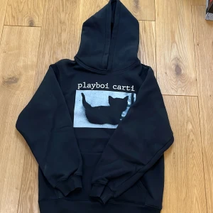 Svart playboy carti hoodie - Säljer en svart hoodie med ett tryck av en katt och texten 'playboi carti' på framsidan. Tröjan har en klassisk design med huva och långa ärmar. Perfekt för en avslappnad stil.