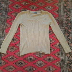 Beige stickad tröja från Ralph Lauren - Säljer en stilren beige stickad tröja från Ralph Lauren med v-ringning och det klassiska logotypbroderiet på bröstet. Tröjan har långa ärmar och en ribbad struktur som ger en elegant look. Perfekt för kyligare dagar.