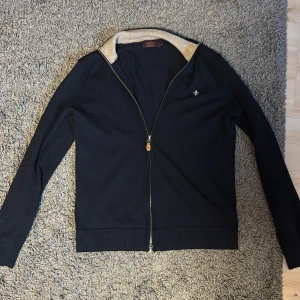 Marinblå kofta från Morris - Snygg marinblå kofta från Morris med dragkedja och en liten broderad detalj på bröstet. Koftan har en kontrasterande krage i beige och långa ärmar. Perfekt för en stilren look.