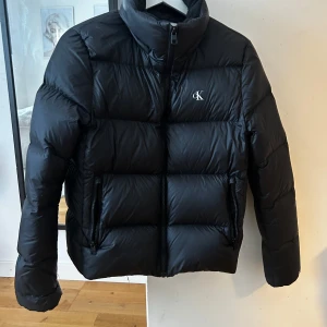 Svart pufferjacka från Calvin Klein Jeans - Säljer en stilren svart pufferjacka från Calvin Klein Jeans. Jackan har en hög krage och dragkedja framtill. Perfekt för kyliga dagar med sin vadderade design. Diskret CK-logga på bröstet.