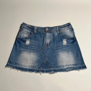 Blå jeanskjol med slitningar - Midja=37,5. Längd=33 cm. Snygg blå jeanskjol med slitningar och fransig kant. Kjolen har en klassisk femficksdesign och en knapp framtill. Perfekt för en avslappnad stil.