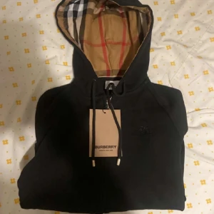 Svart huvtröja från Burberry - Snygg svart huvtröja från Burberry med klassiskt rutigt mönster i huvan. Tröjan har dragkedja framtill och ribbade muddar vid ärmslut och nederkant. Perfekt för en stilren look.