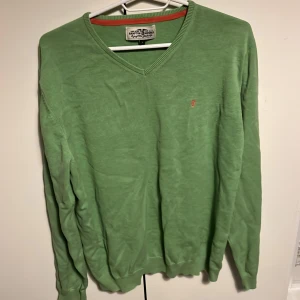 Grön tröja från Hampton Republic - Säljer en grön tröja från Hampton Republic med v-ringning och långa ärmar. Tröjan har en liten logga på bröstet och ribbade kanter. Perfekt för en avslappnad stil.