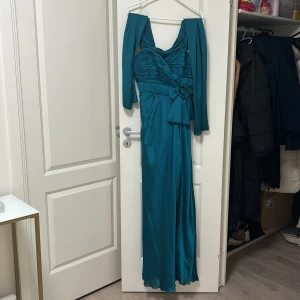 Turkos långklänning med avtagbara ärmar - Elegant turkos långklänning med avtagbara ärmar och en vacker blomdetalj i midjan. Klänningen har en omlott-design och smala axelband, vilket ger en stilren look. Perfekt för speciella tillfällen.