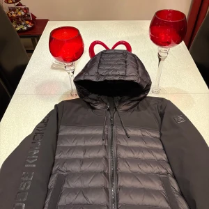 Svart pufferjacka från Moose Knuckles - Snygg svart pufferjacka från Moose Knuckles med huva och dragkedja. Jackan har ett quiltat mönster och logotyp på ärmen. Perfekt för kyliga dagar.