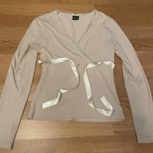 Beige omlottopp från Zentex - Säljer en elegant beige omlottopp från Zentex med långa ärmar och spetsdetaljer vid halsen. Toppen har ett knytband i satin som ger en fin passform. Perfekt för en stilren look.