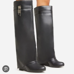 Svarta höga stövlar med låsdetalj - Eleganta svarta boots i skinn med en unik låsdetalj på sidan. De har en spetsig tå och en diskret klack, vilket ger en stilren och modern look. Köpta second hand, använda en gång. Påminner om Givenchy bootsen