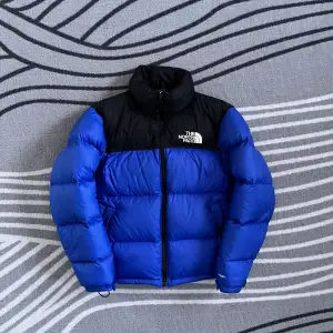 Säljer en snygg blå och svart dunjacka från The North Face. Jackan har en dragkedja framtill och är perfekt för kalla dagar. Den är långärmad och har en klassisk pufferdesign med märkets logga på bröstet. Den passar som en s/m