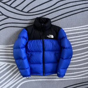 Blå och svart dunjacka från The North Face - Säljer en snygg blå och svart dunjacka från The North Face. Jackan har en dragkedja framtill och är perfekt för kalla dagar. Den är långärmad och har en klassisk pufferdesign med märkets logga på bröstet. Den passar som en s/m