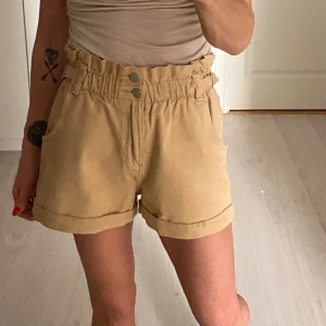 Beige shorts med hög midja - Snygga beige shorts med hög midja och justerbara spännen. De har två knappar framtill och uppvikta benslut för en avslappnad look. Perfekta för varma dagar!